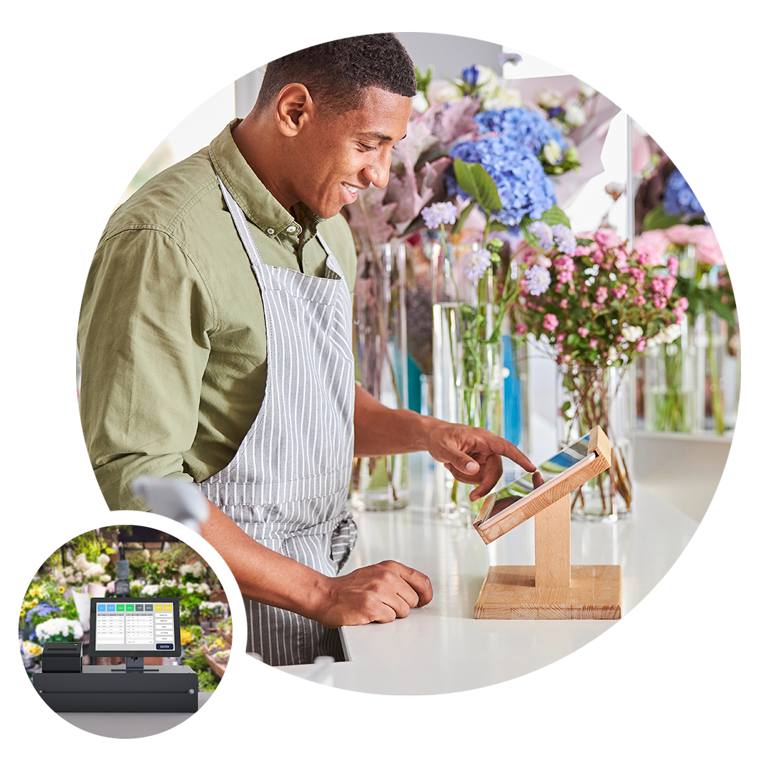 A person using a tablet at a flower shop counter with a point-of-sale system in the foreground. A person using a tablet at a flower shop counter with a point-of-sale system in the foreground.