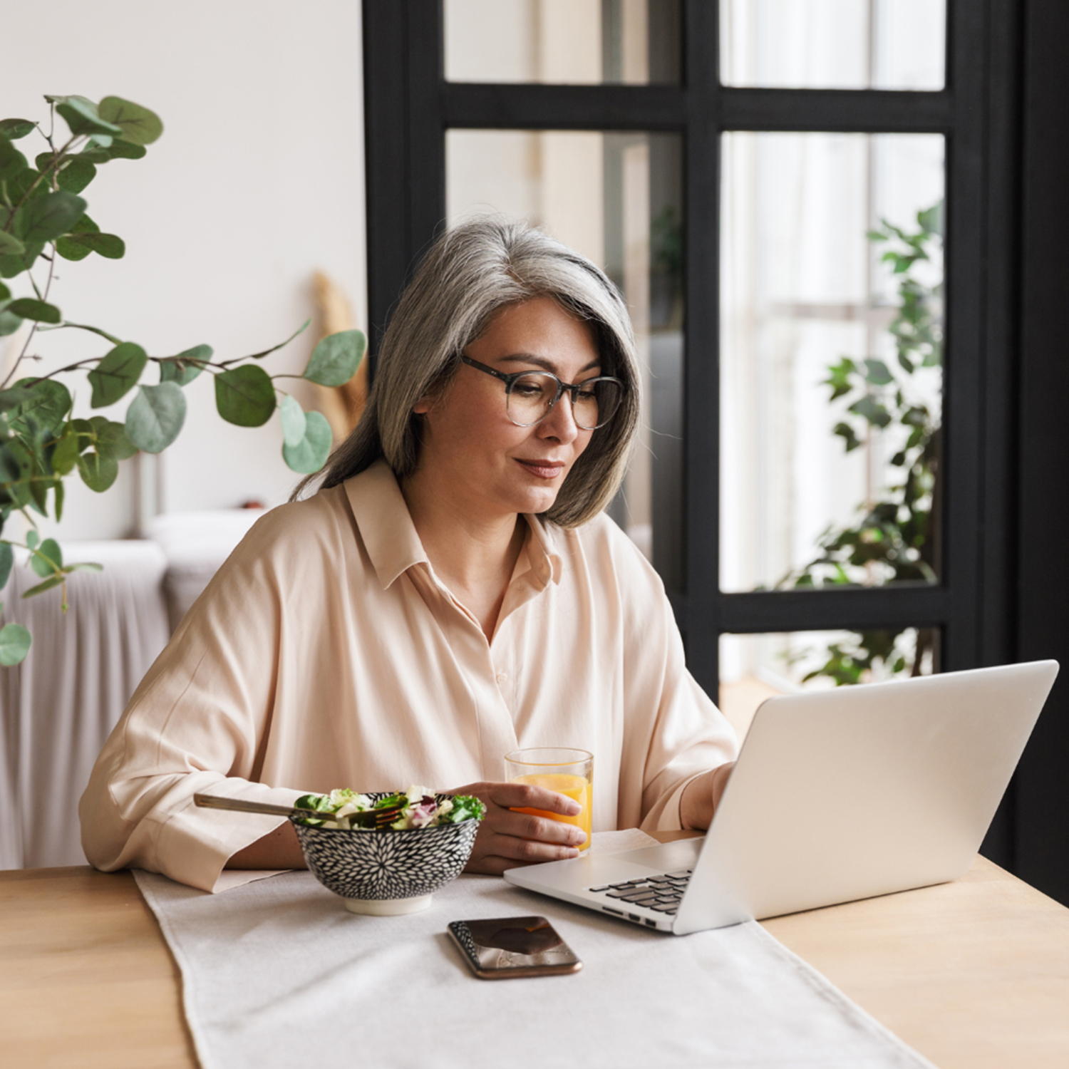 Woman using laptop at kitchen table, holding orange juice; salad and phone nearby, plants in background.