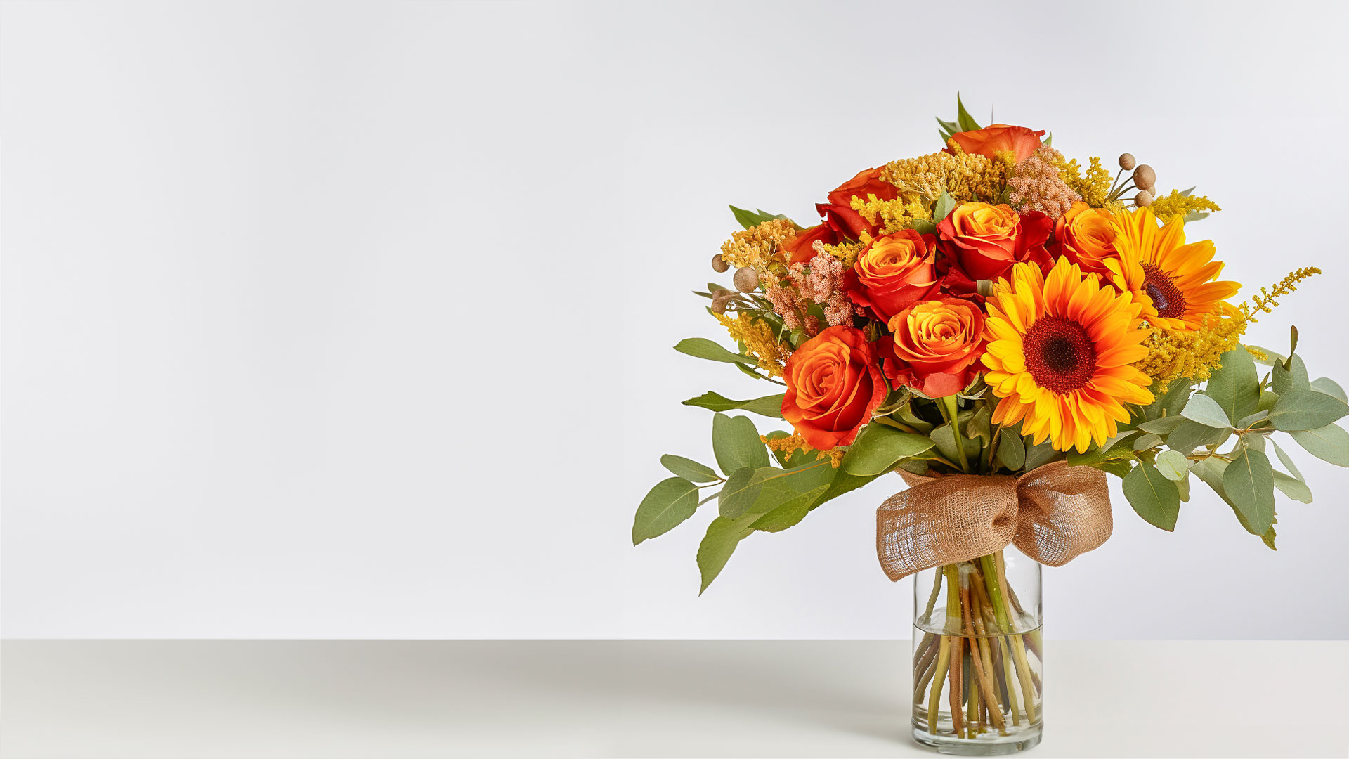 Nebula Fashion banner featuring autumn arrangement of yellow sunflowers, orange roses, and greenery tied with burlap in a clear vase on a plain surface.
