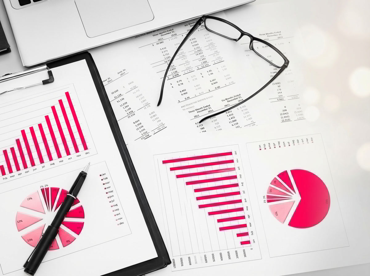 Overhead view of reports with red bar and pie charts beside a laptop, glasses, clipboard, and black pen.